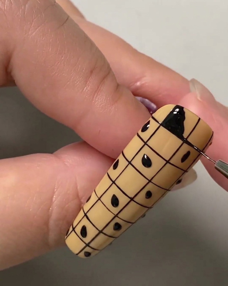Primer plano de un pincel liner rellenando el cuadrado superior izquierdo de una cuadrícula en una uña nude con esmalte en gel negro.