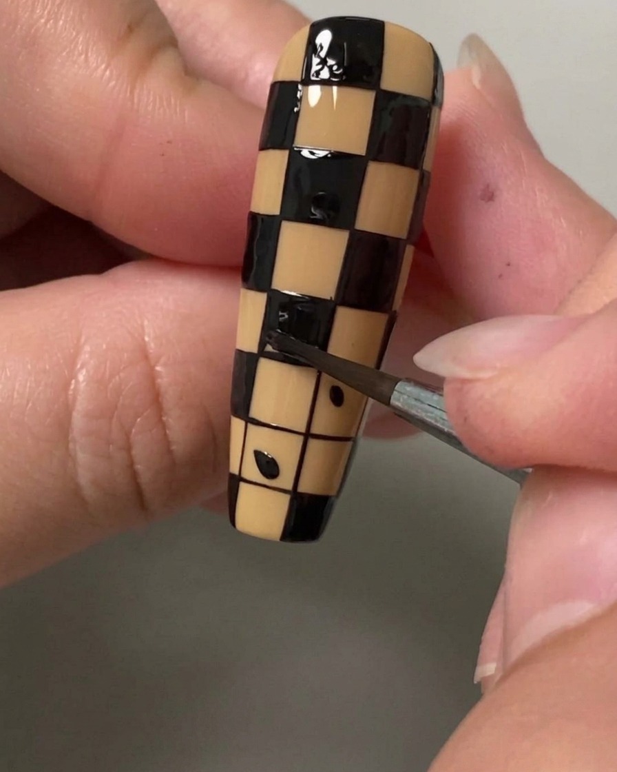 Primer plano de un pincel de detalle fino rellenando un cuadrado pequeño con esmalte negro para crear un patrón de tablero de ajedrez en una uña.