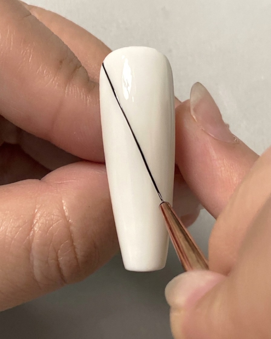 Primer plano de un pincel liner fino dibujando una línea diagonal negra larga a través de una uña blanca.