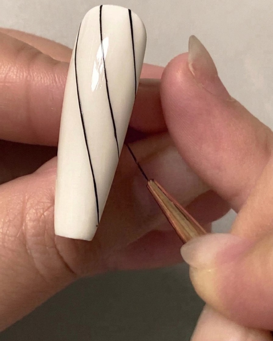 Primer plano de un pincel de nail art fino dibujando una segunda línea diagonal negra paralela en la mitad superior de una uña blanca.