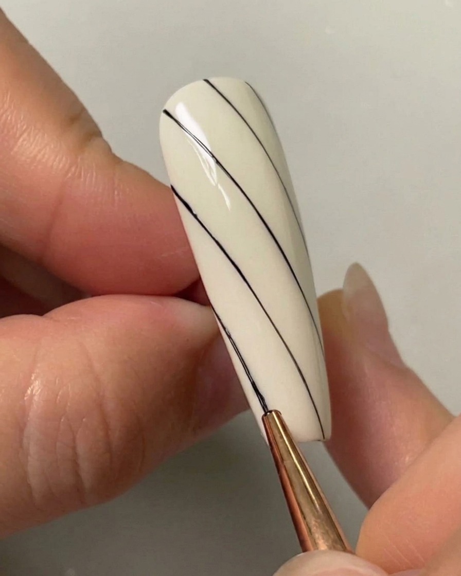 Un pincel de nail art dibujando una tercera línea diagonal negra en el lado inferior izquierdo de una uña pintada de blanco.