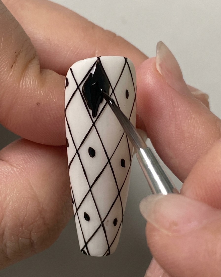 Un pincel liner fino rellenando cuidadosamente la forma de diamante superior con esmalte en gel negro sobre una uña con cuadrícula blanca previamente dibujada.