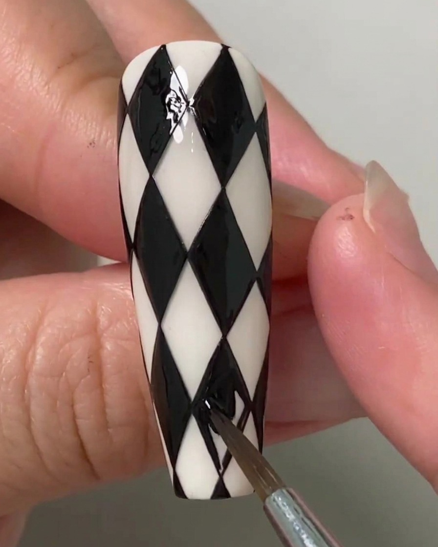 Una artista de uñas usando un pincel fino para rellenar las últimas formas de diamante restantes en una uña con patrón argyle en blanco y negro.
