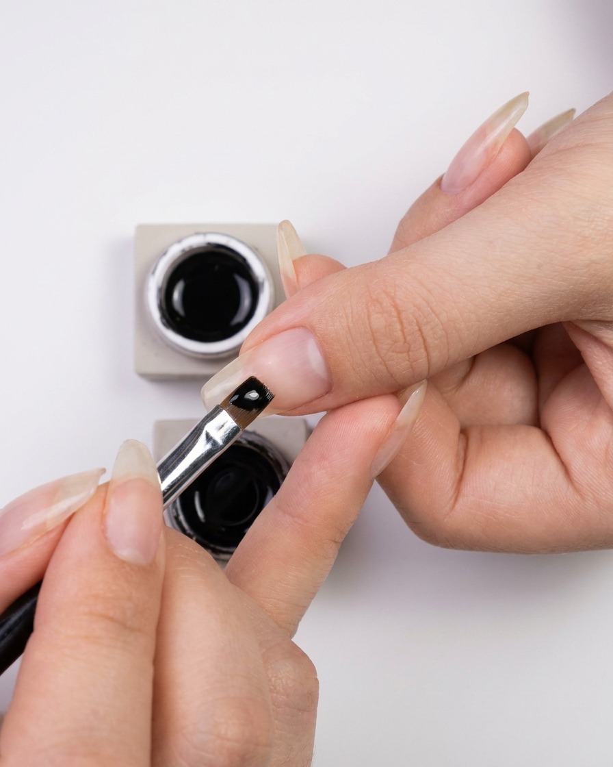Mano sumergiendo un pincel fino de nail art en un pequeño tarro de esmalte en gel negro brillante.