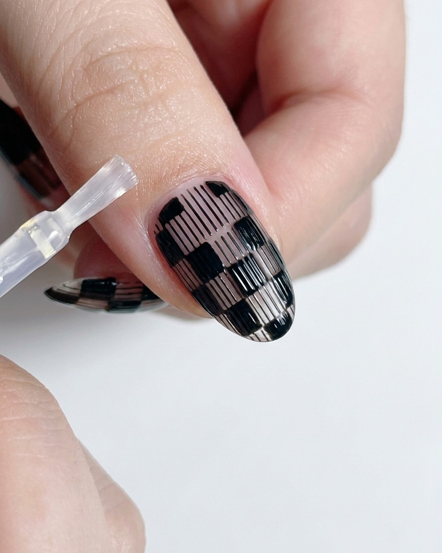 Pincel aplicando esmalte transparente y brillante sobre un diseño de uñas a cuadros en blanco y negro.