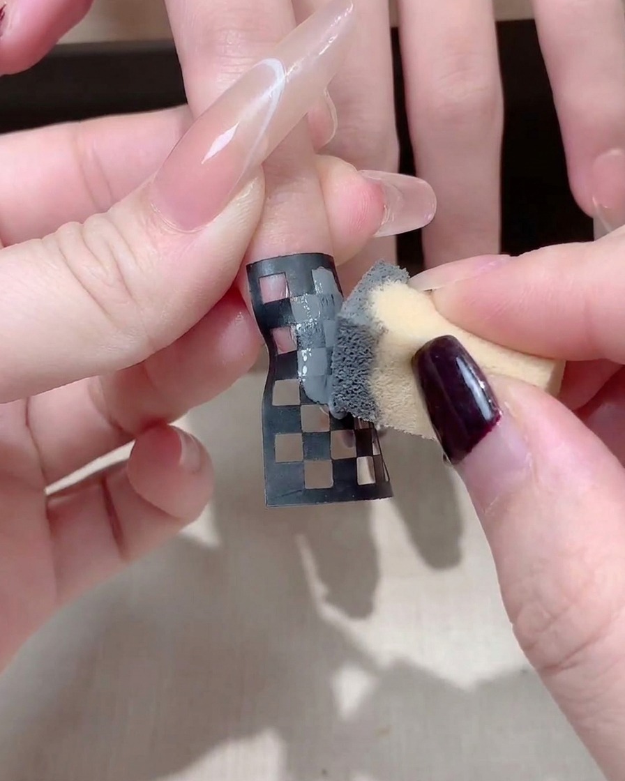 Un primer plano de una manicurista ajustando una pegatina de uñas a cuadros blancos y negros en una uña larga y cuadrada.