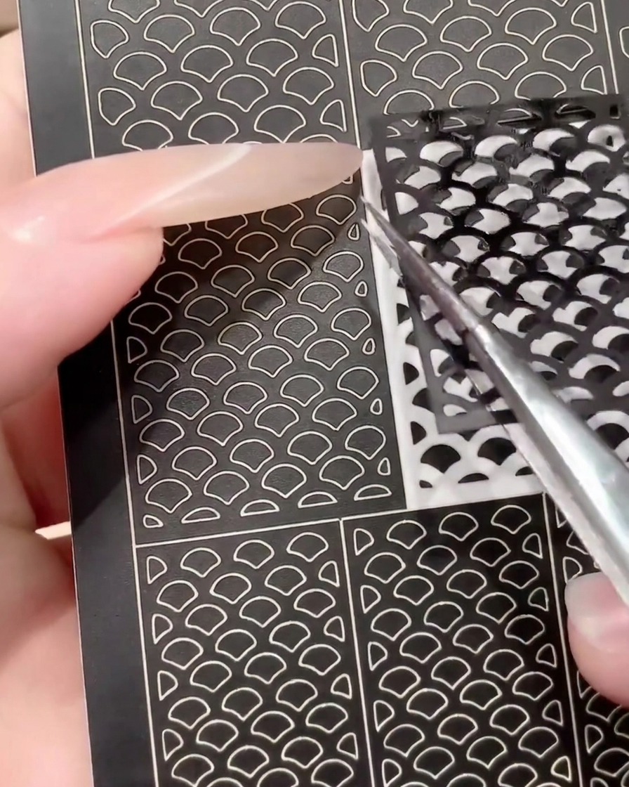 Un par de pinzas despegando una hoja de pegatinas de nail art negra usada para mostrar su reutilización.