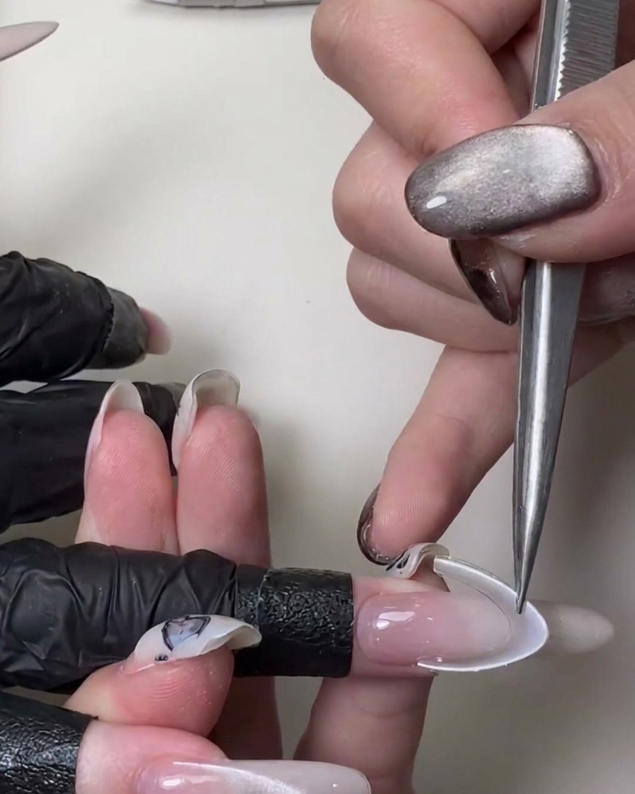 Primer plano de una mano usando unas pinzas de metal para aplicar una pegatina blanca curva de sonrisa francesa sobre una uña larga de color rosa translúcido.