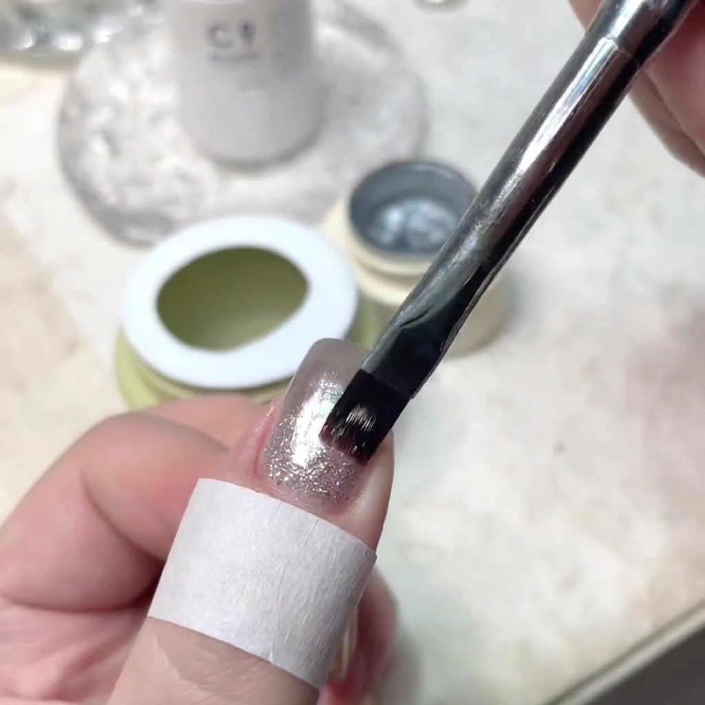 Un pincel de uñas aplicando con precisión un trazo fino de gel brillante en el centro de una uña manicurada.