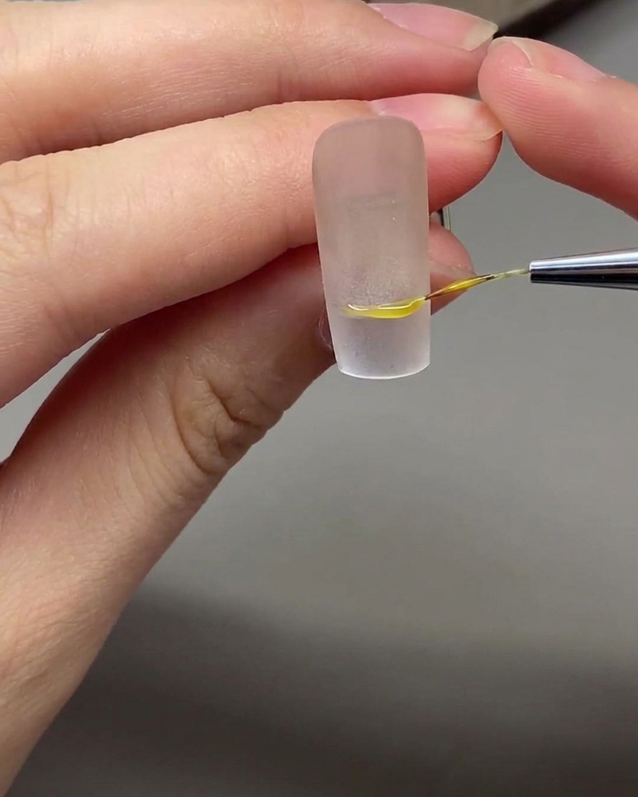 Un pincel fino para nail art dibujando una línea de sonrisa francesa precisa sobre una base de uña nude.