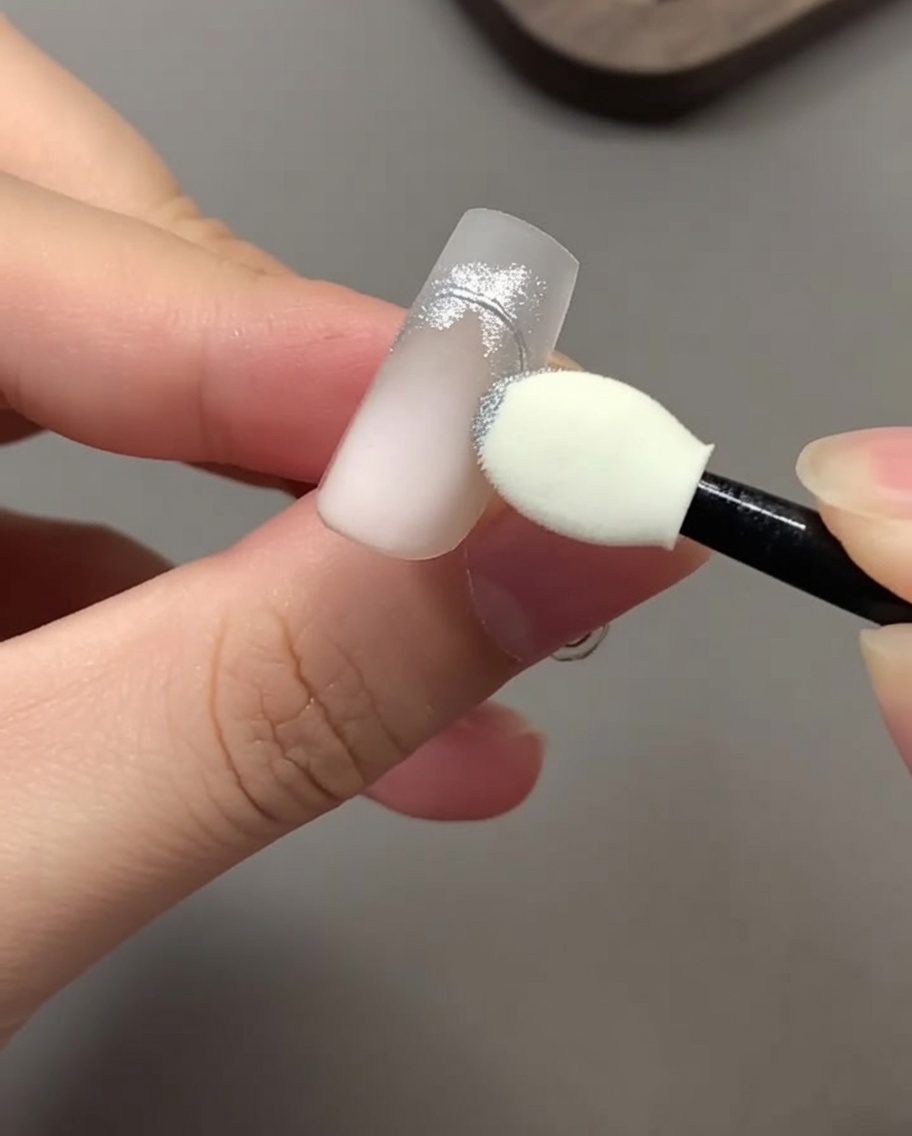 Un aplicador de esponja para sombras frotando polvo cromado plateado sobre un diseño de uñas.