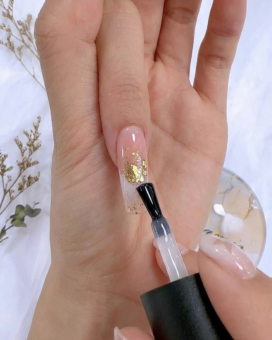 Un pincel de esmalte aplicando un top coat transparente sobre una uña decorada con detalles en pan de oro.