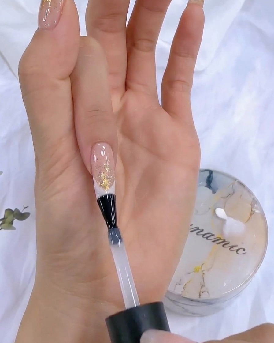 Una mano sosteniendo un frasco de top coat cerca de una lámpara de uñas UV/LED durante la etapa final de curado.