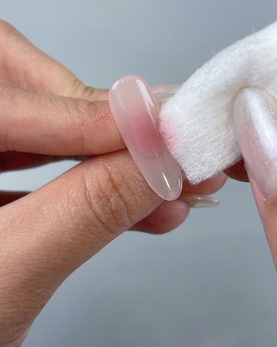 Una mano usando un bloque de esponja blanco para aplicar esmalte en gel rosa suave en el centro de una uña postiza transparente con forma almendrada.