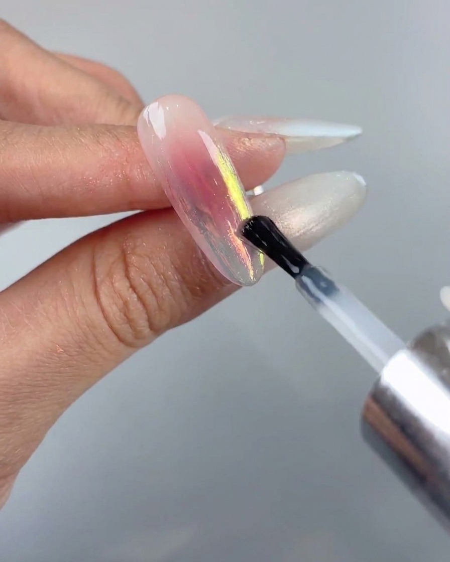 Un pincel de uñas aplicando una capa final de sellado con top coat suave sobre un diseño de uña cromado aurora.