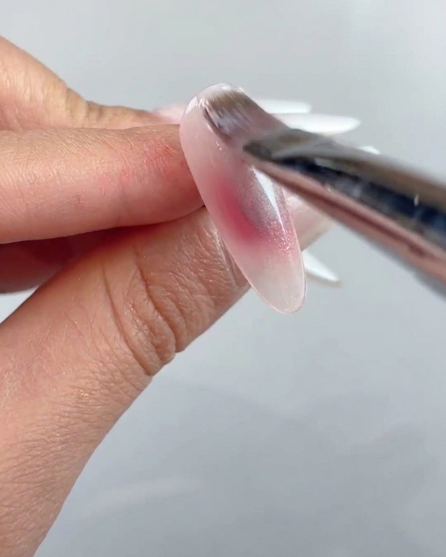 Un pincel de uñas aplicando una capa de gel ojo de gato transparente sobre toda la superficie de la uña preparada.