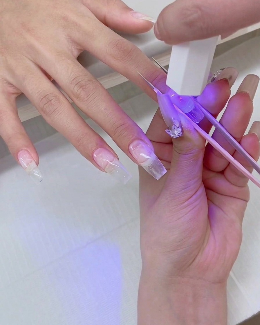 Primer plano de una mano sosteniendo una mini lámpara UV sobre una extensión de uña transparente mientras unas pinzas estabilizan el tip.