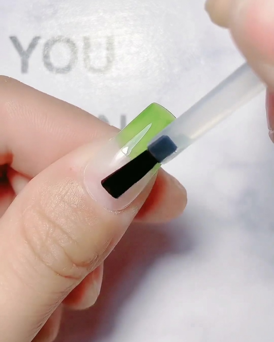 Un pincel de uñas aplicando gel verde oliva en la mitad superior de una uña natural.