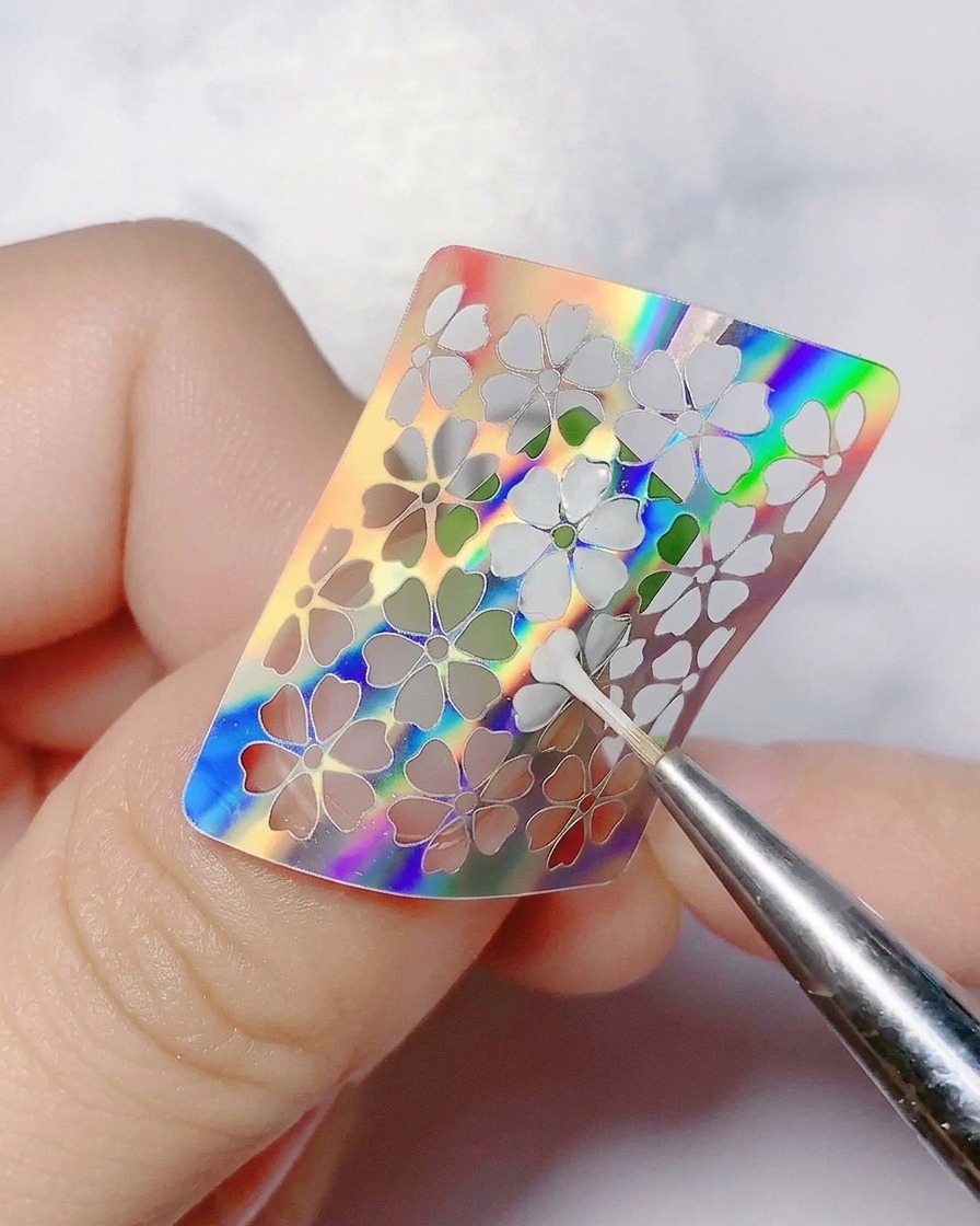Una herramienta fina retirando una pegatina de flor holográfica de la uña.