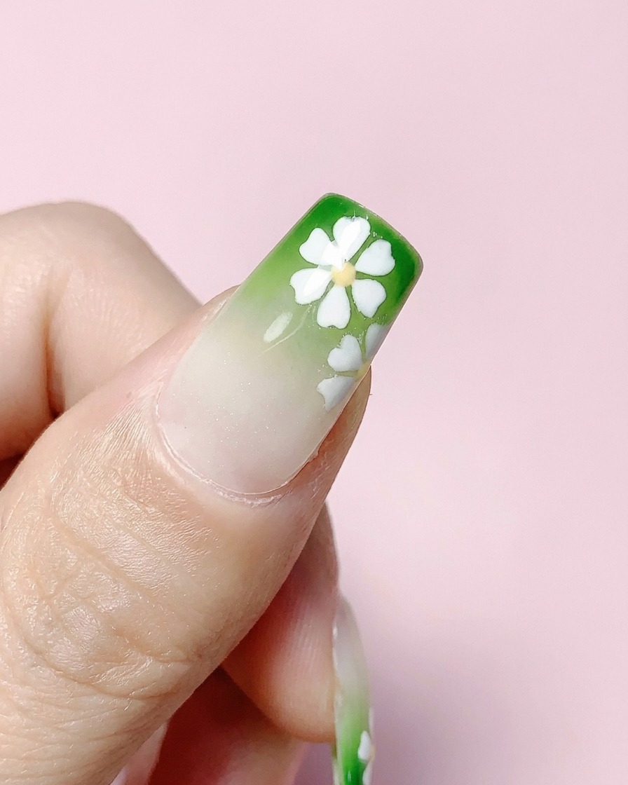 Una uña cuadrada terminada con degradado verde y flores blancas siendo cubierta con una capa superior transparente.