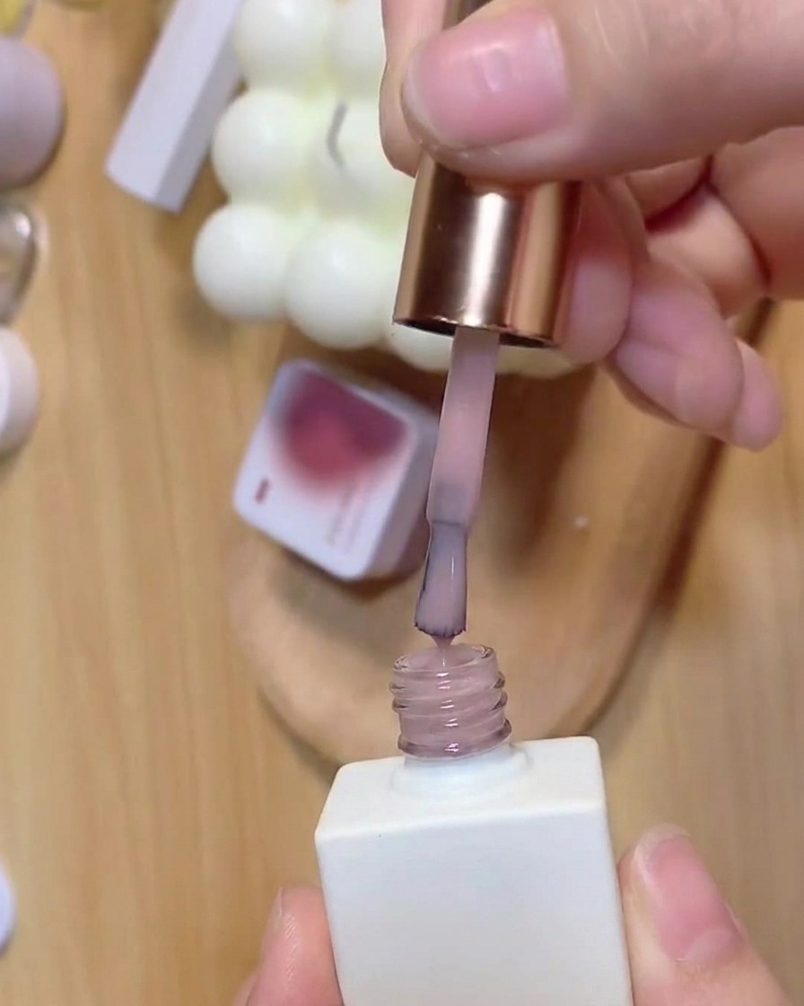 Un pincel extrayendo capa base rosa translúcida de un frasco de esmalte de uñas blanco.