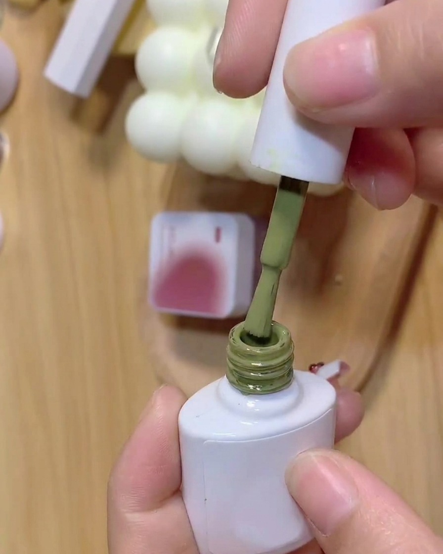 Un pincel sumergiéndose en un frasco de esmalte de gel verde con un frasco de gel rosa de fondo.