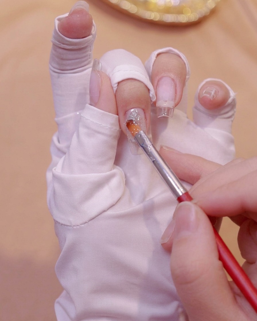 Un pincel fino aplicando gel con purpurina plateada cerca de la base de una extensión de uña transparente en una mano con guante.