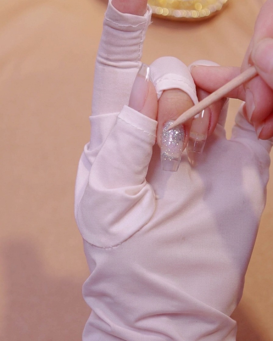 Un palito de naranjo presionando suavemente lentejuelas de mariposa incrustadas en gel con purpurina sobre una uña.
