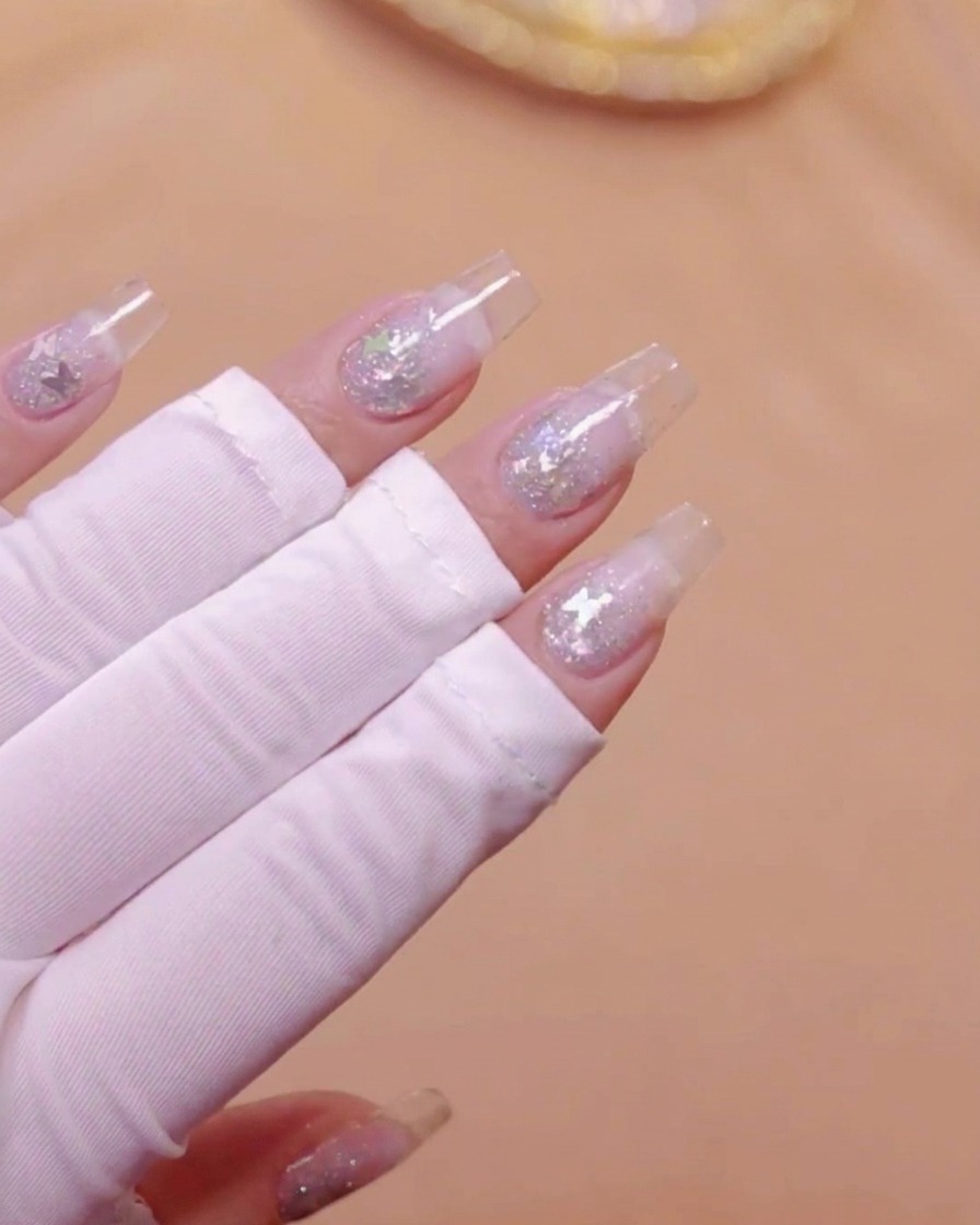 Una mano con extensiones de uñas transparentes y purpurina de mariposa incrustada colocada bajo una lámpara UV.