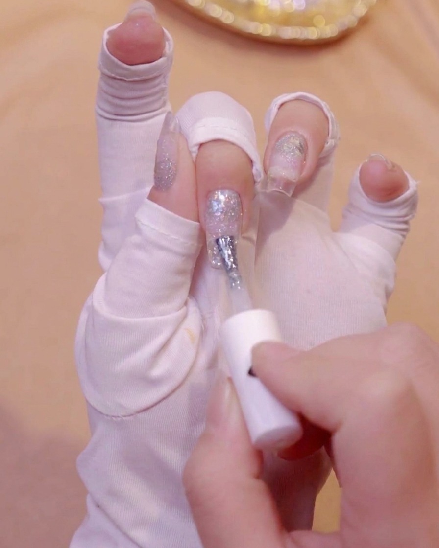 Una mano con guante blanco usando un pincel para aplicar esmalte en gel blanco lechoso en la punta de una uña.