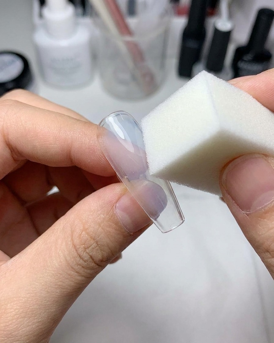 Una pequeña esponja cosmética presionando gel azul claro en el centro de una uña postiza transparente.