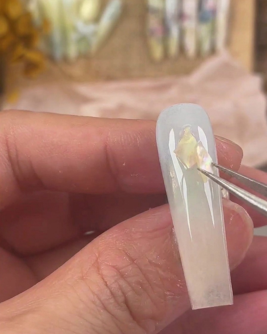 Unas pinzas finas colocando piezas de concha iridiscente sobre la superficie húmeda de la uña.