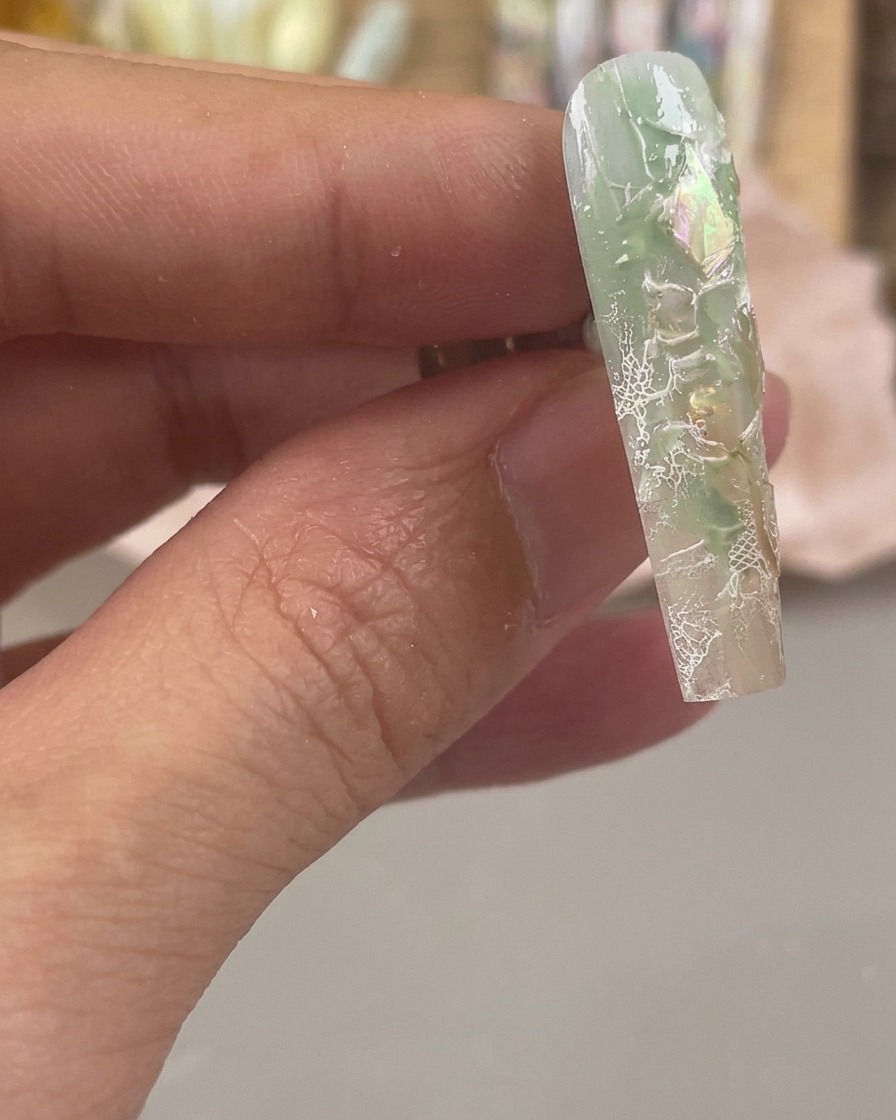 Primer plano de una uña en tonos verdes y transparentes con escamas de concha iridiscente y patrones de encaje blanco estampados.