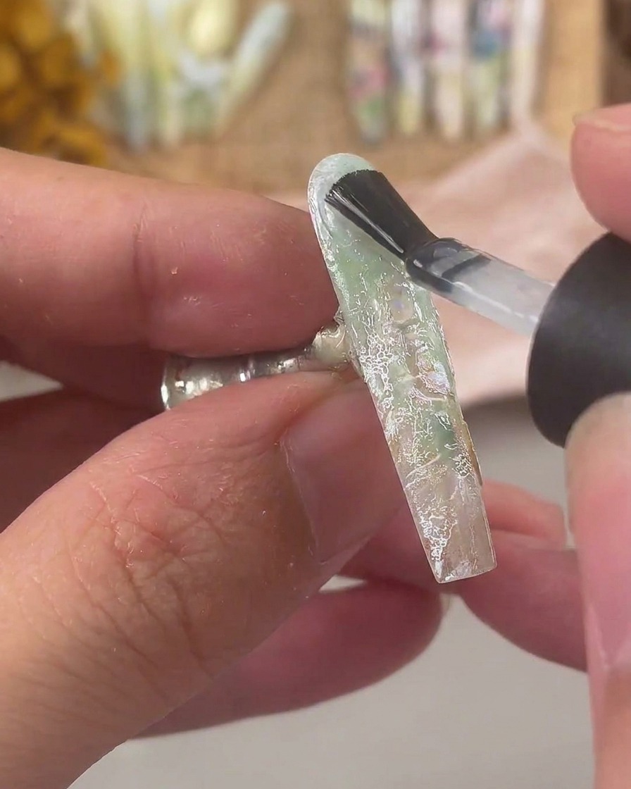 Un pincel aplicando una capa gruesa de gel constructor transparente sobre una uña decorada con encaje y escamas de concha.