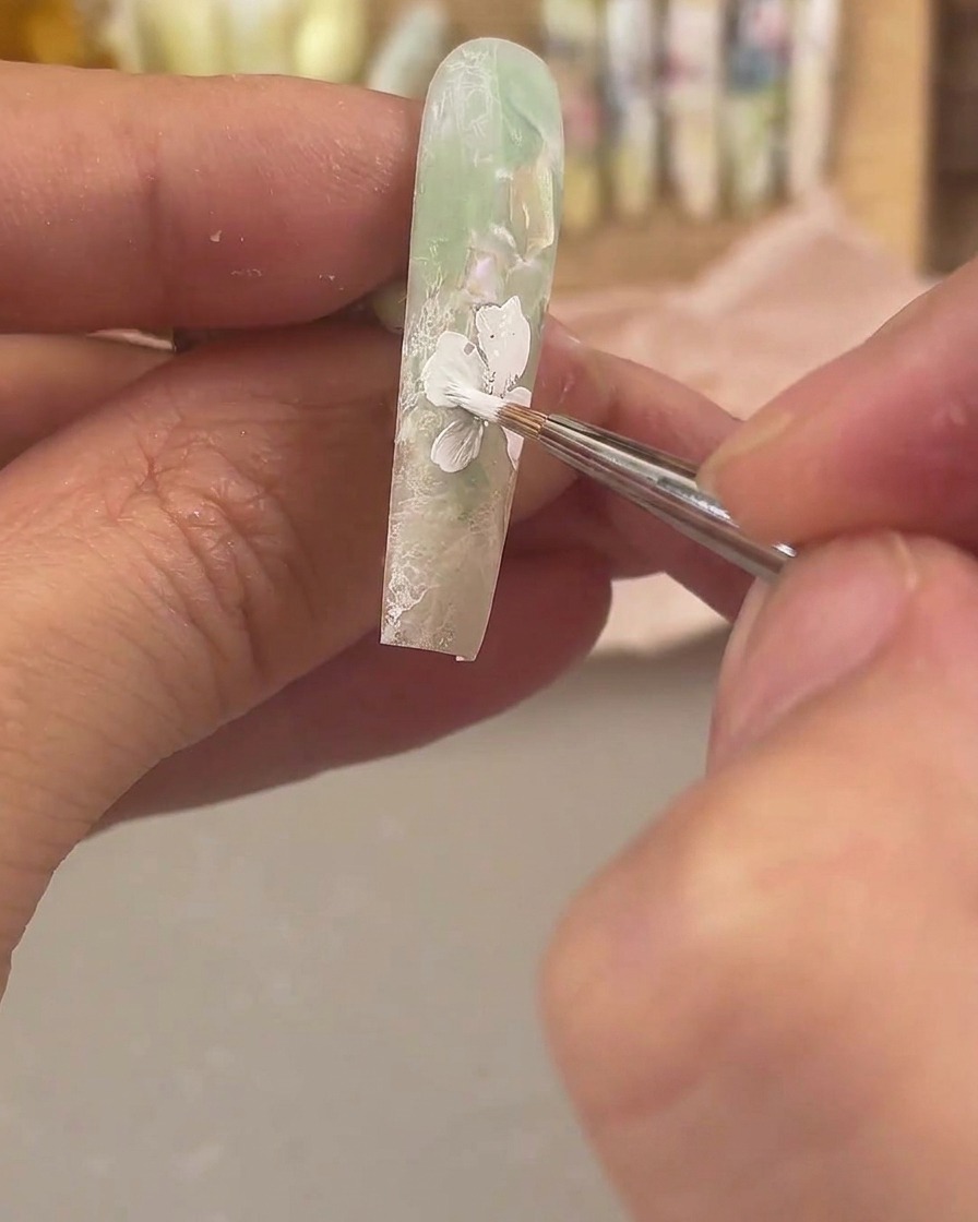 Un pincel de detalle fino aplicando gel de pintura blanco para dibujar una flor grande sobre una uña verde mate.