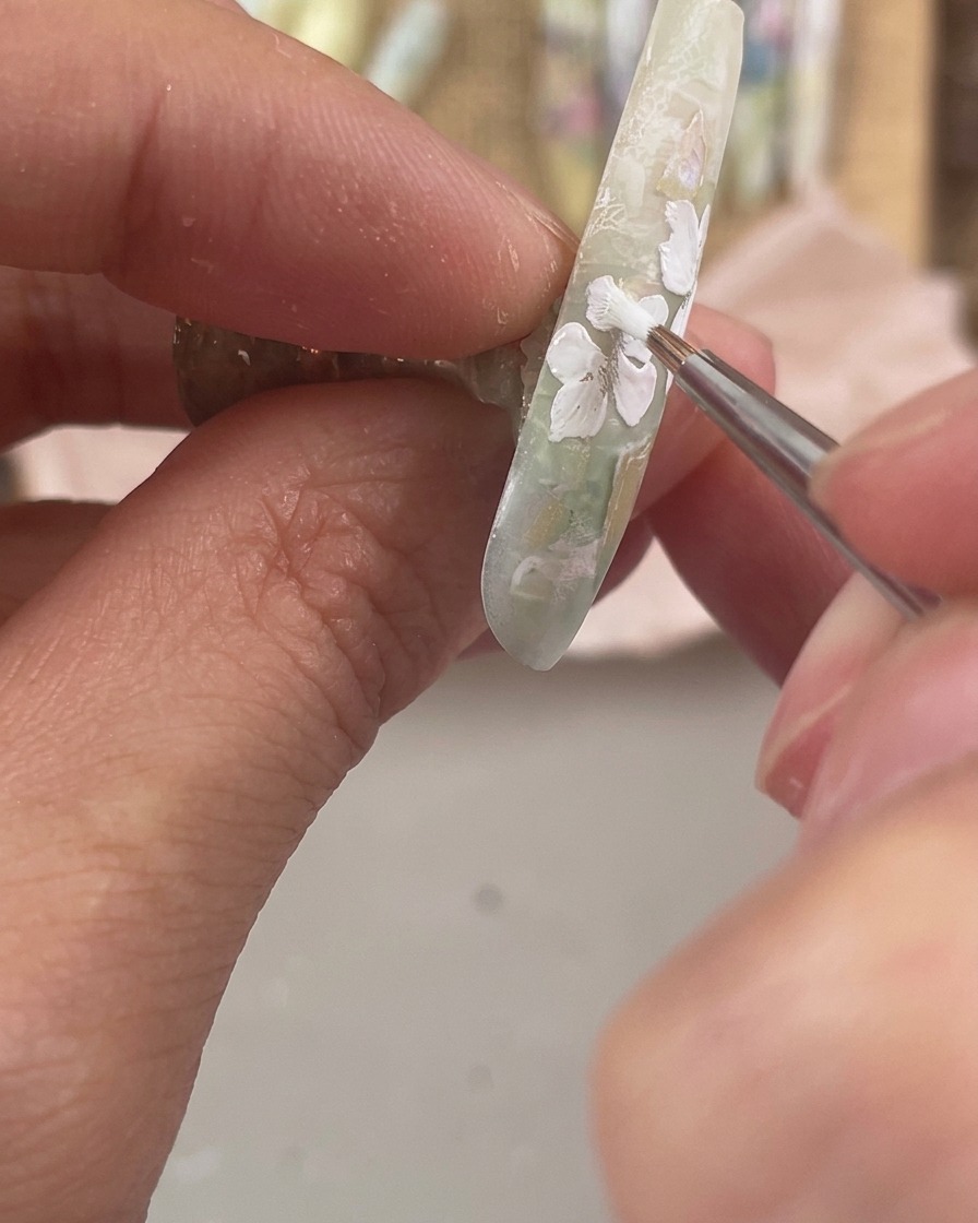 Un pincel de detalle fino pintando tres pequeños pétalos blancos a lo largo del borde inferior de una uña decorada.