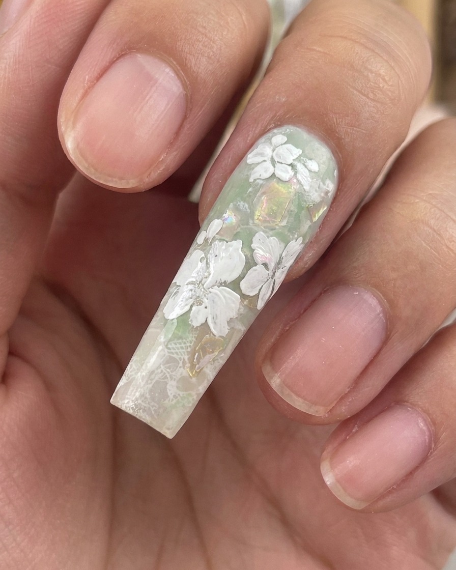 Primer plano de una uña esculpida y larga que muestra flores y pequeñas hojas de gel blanco terminadas sobre un fondo verde con conchas.