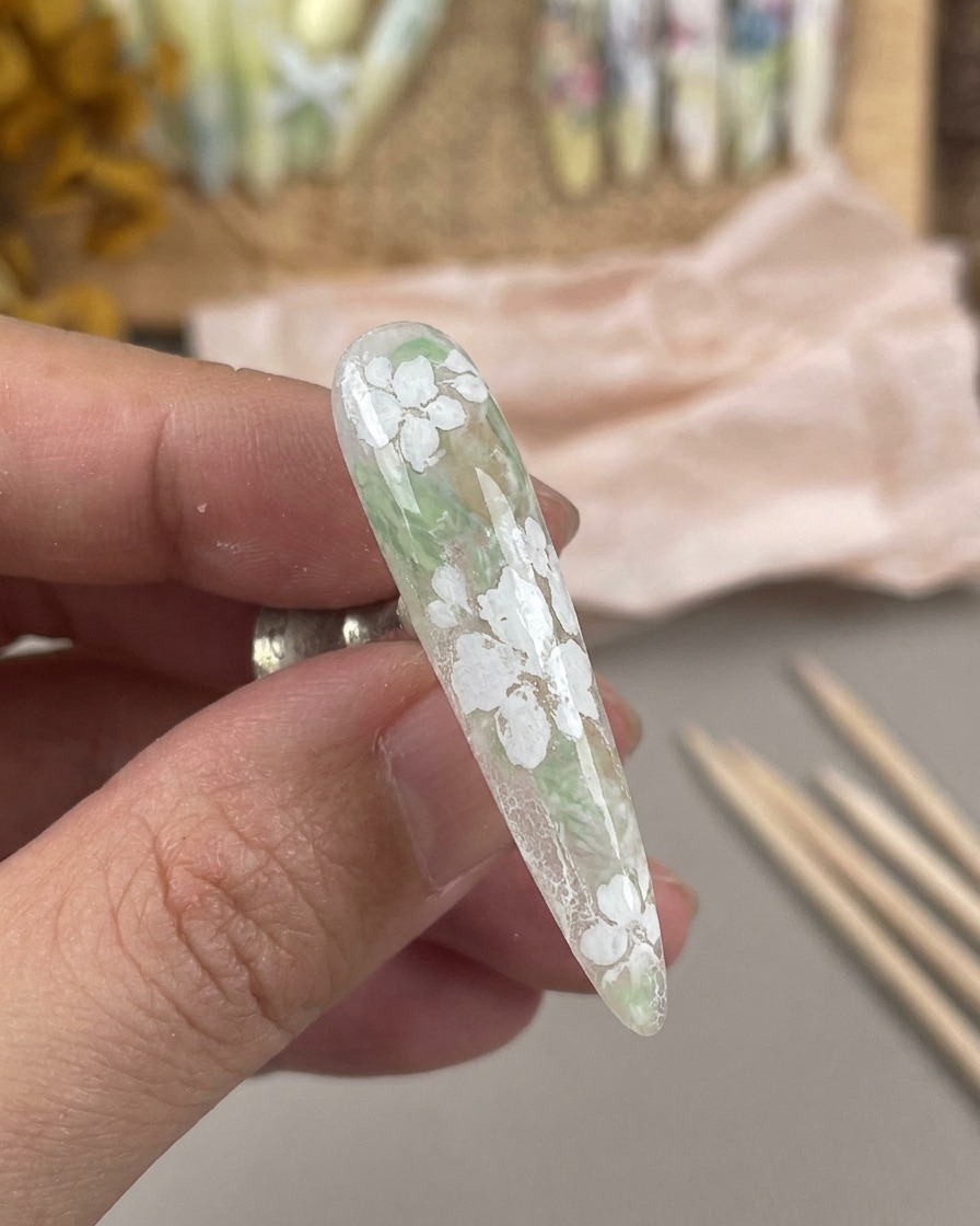 Una uña larga con acabado mate que presenta un diseño floral blanco, sostenida en un soporte magnético para uñas.