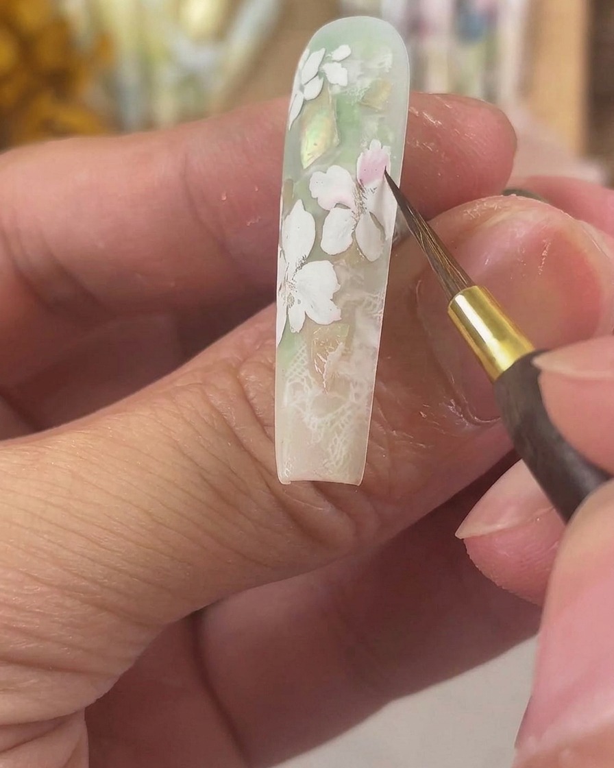Un pincel fino aplicando pigmento de acuarela rosa translúcido sobre pétalos de gel blanco mate en una uña larga esculpida.