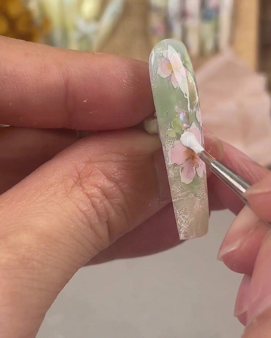 Un pincel delineador aplicando gel de pintura blanco para formar una capa extra de pétalos en un diseño de nail art floral rosa.