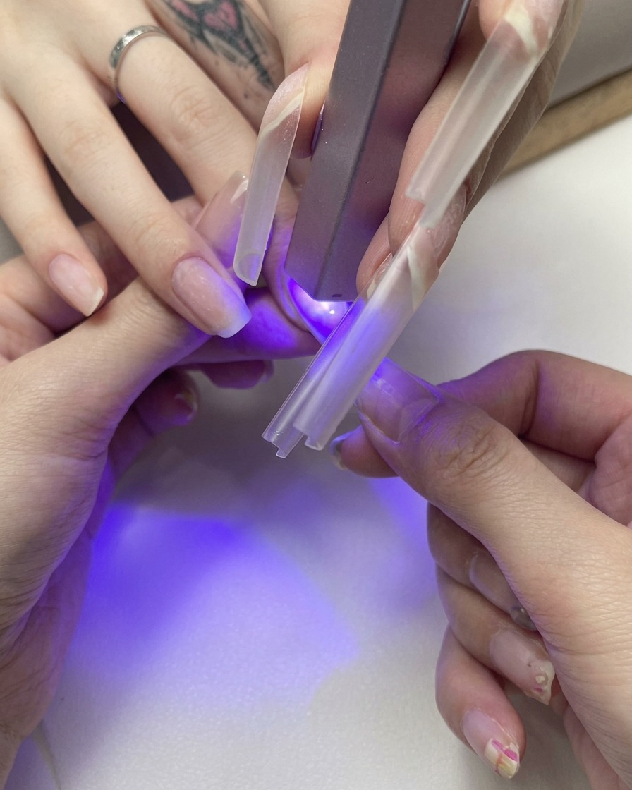Mano sosteniendo un tip de extensión transparente sobre una uña natural bajo una pequeña luz UV violeta.