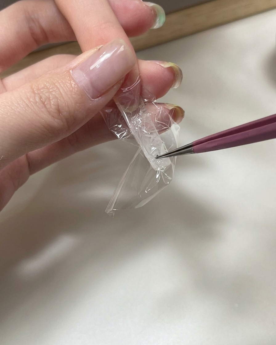 Pinzas presionando un trozo de papel film transparente en la parte inferior de una extensión de uña para crear arrugas.
