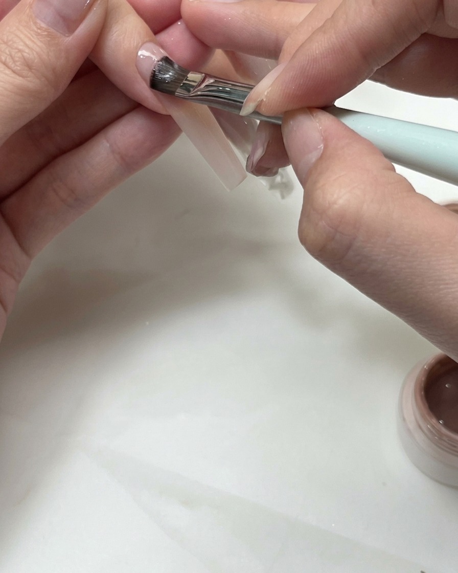 Pincel plano aplicando gel nude para formar una línea de sonrisa francesa en una uña.