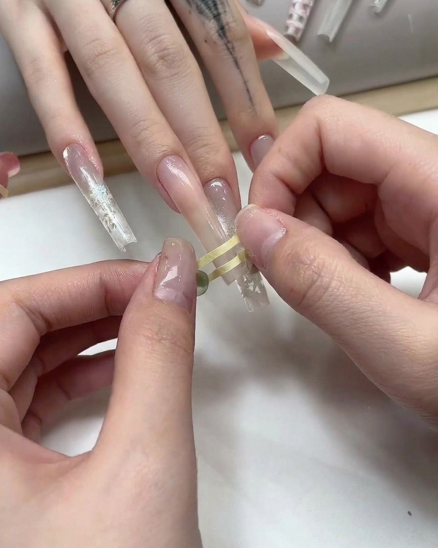 Dedos colocando cinta fina dorada en un patrón de cuadrícula sobre una uña de gel nude.