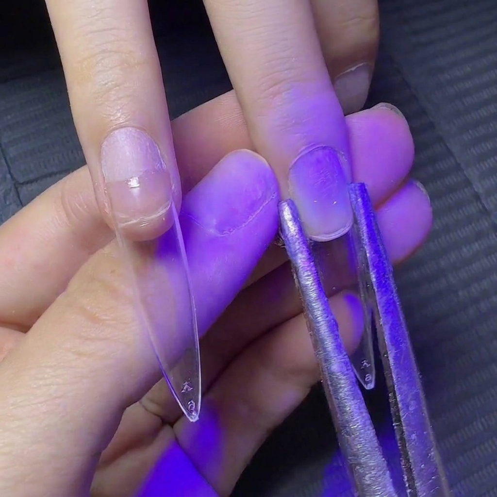 Pinzas de metal sujetando los lados de una extensión de uña transparente bajo luz UV para dar forma a la curva.