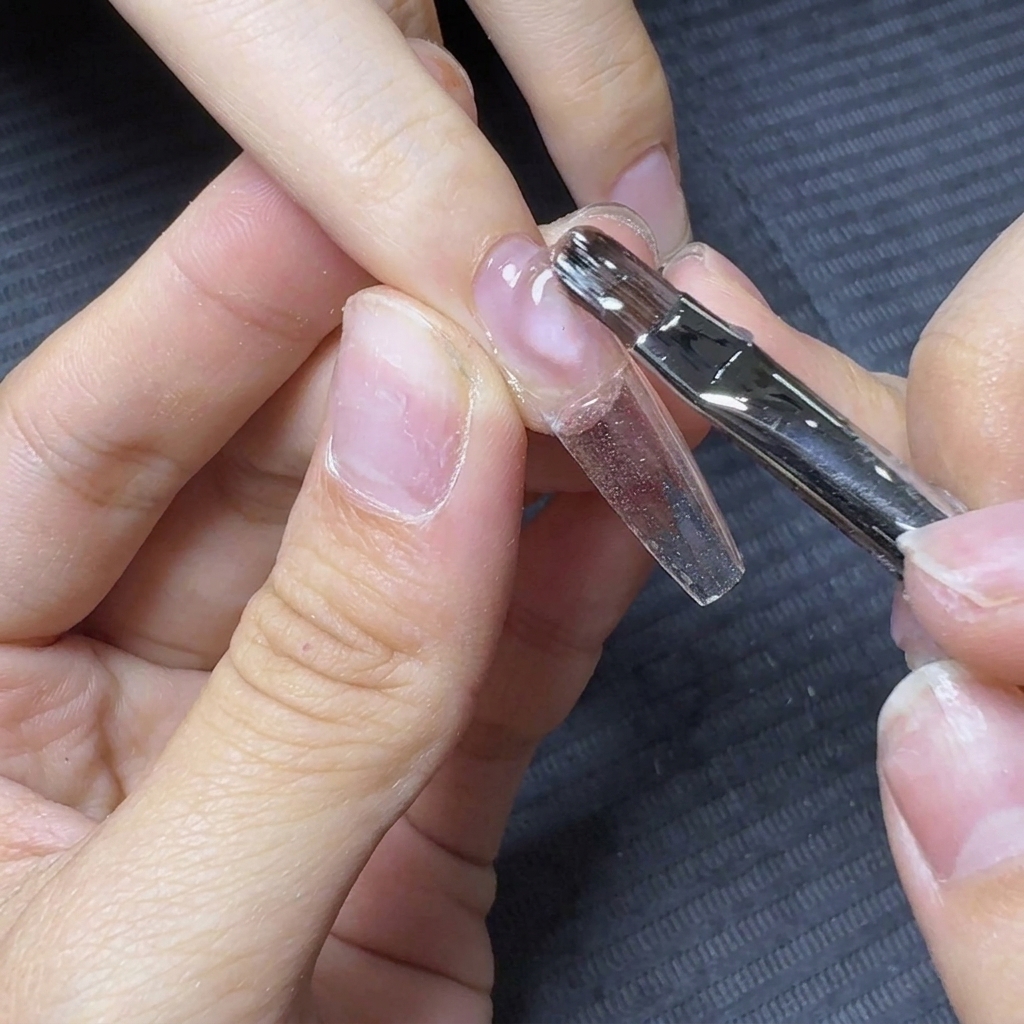Un pincel de uñas flotando y arrastrando gel constructor transparente a lo largo de un tip de extensión.