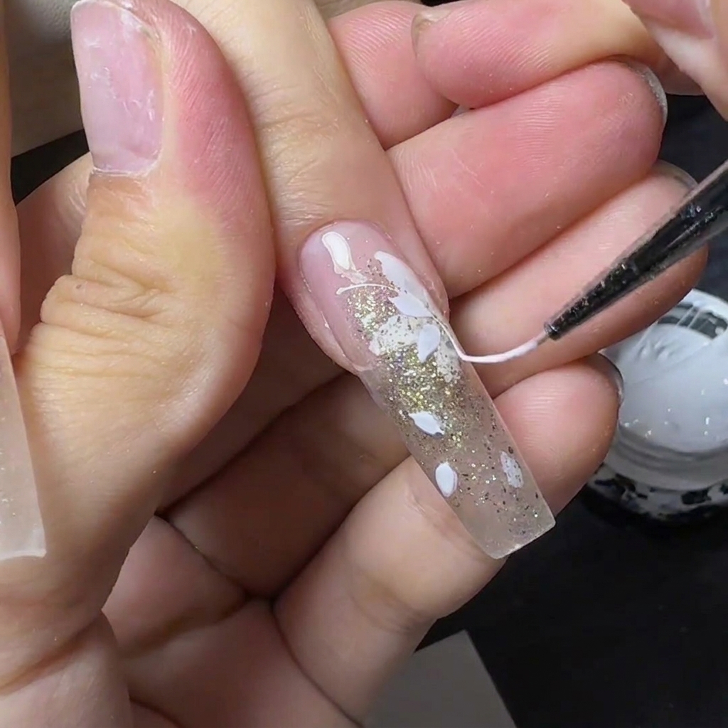 Una herramienta fina tirando de spider gel blanco en forma de hilo a través de una uña decorada con flores y purpurina.
