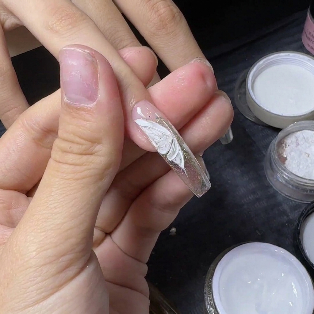 El pulgar de un manicurista sosteniendo una uña para fijar un pequeño cristal plateado en una mariposa blanca pintada.