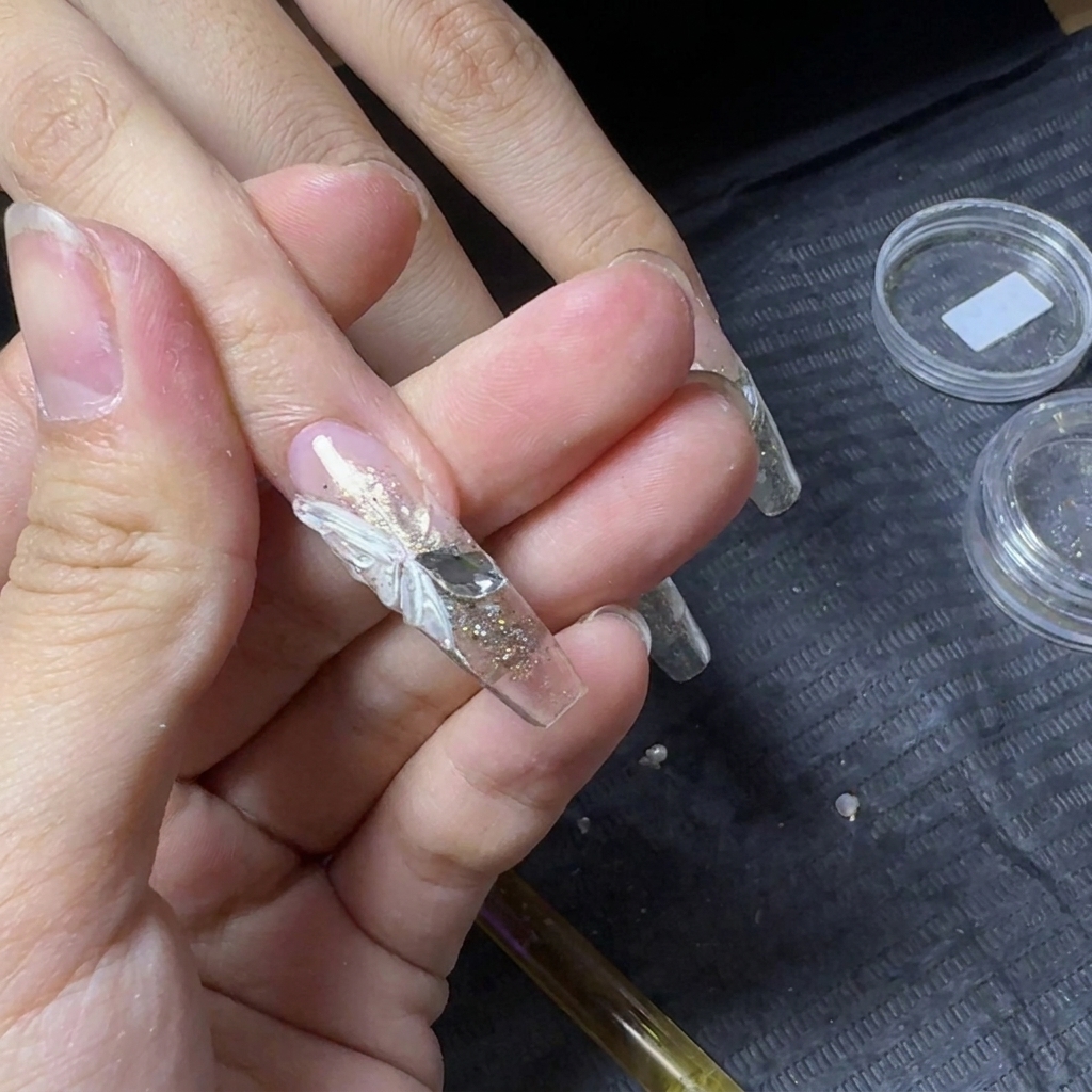 Primer plano de una extensión de uña transparente con un ala de mariposa blanca en 3D y dos cristales colocados al lado.