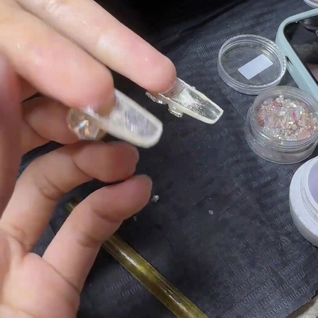 Una mano sosteniendo una extensión de uña transparente terminada boca abajo, exponiendo la parte inferior para aplicar top coat.